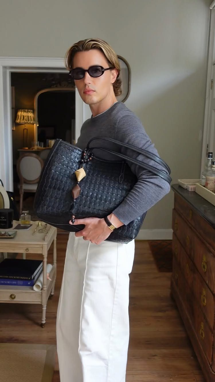 A man in a grey shirt and white trousers carrying a large dark leather tote bag.