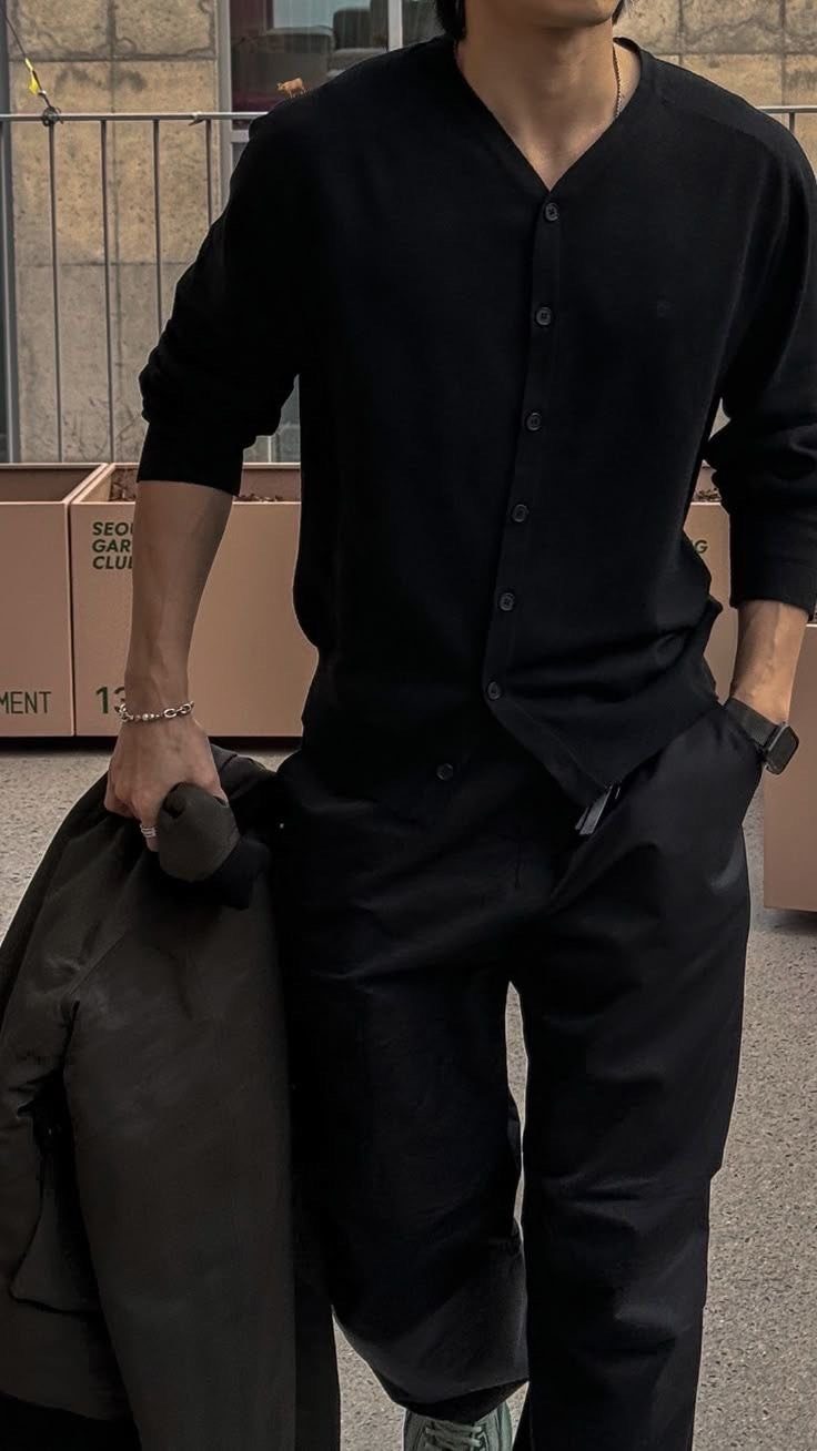 A man dressed in a black button-up cardigan and black trousers holding an olive jacket.