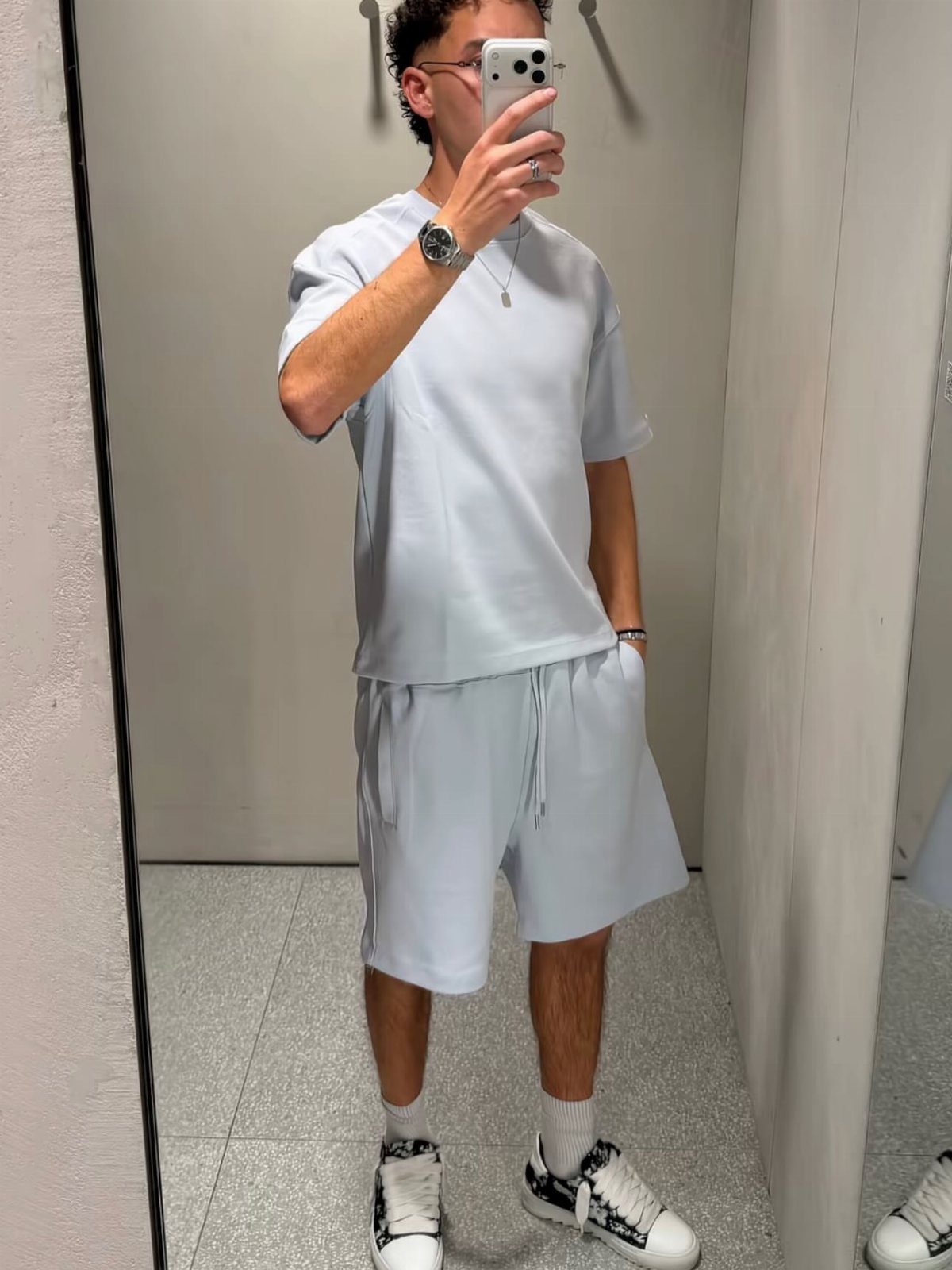 Man in a light blue monochromatic t-shirt and shorts outfit taking a mirror selfie.