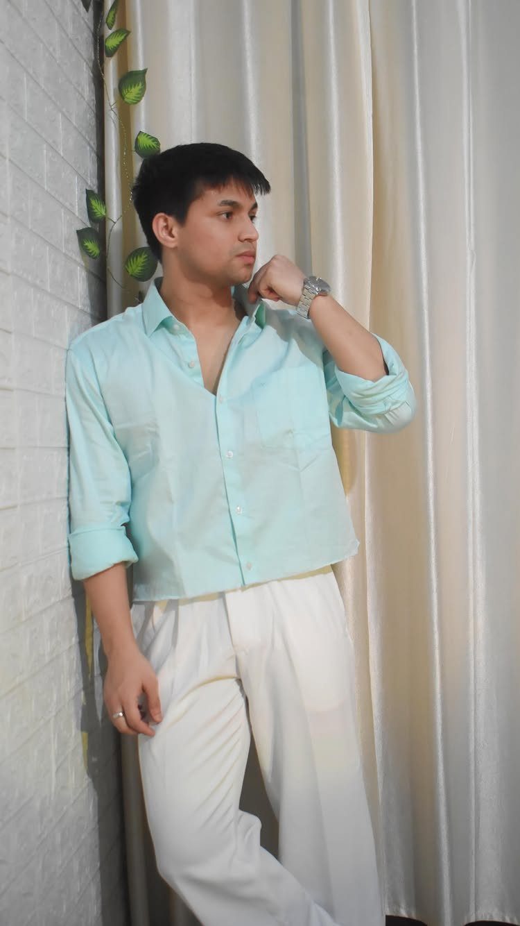 A young man in a turquoise shirt and white pants leaning against a white brick wall.