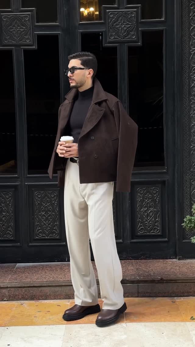 A man in a brown jacket and cream trousers standing before a black door.