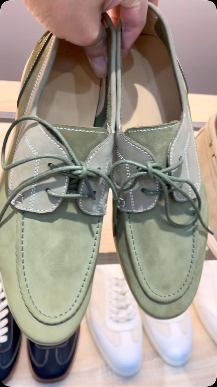 Sage green suede boat loafers with thin laces being held by a hand.