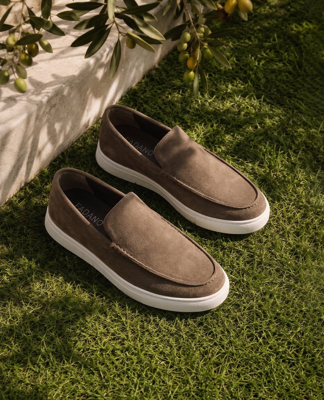 Brown suede slip-on loafers with white soles sitting on green grass near olive branches.
