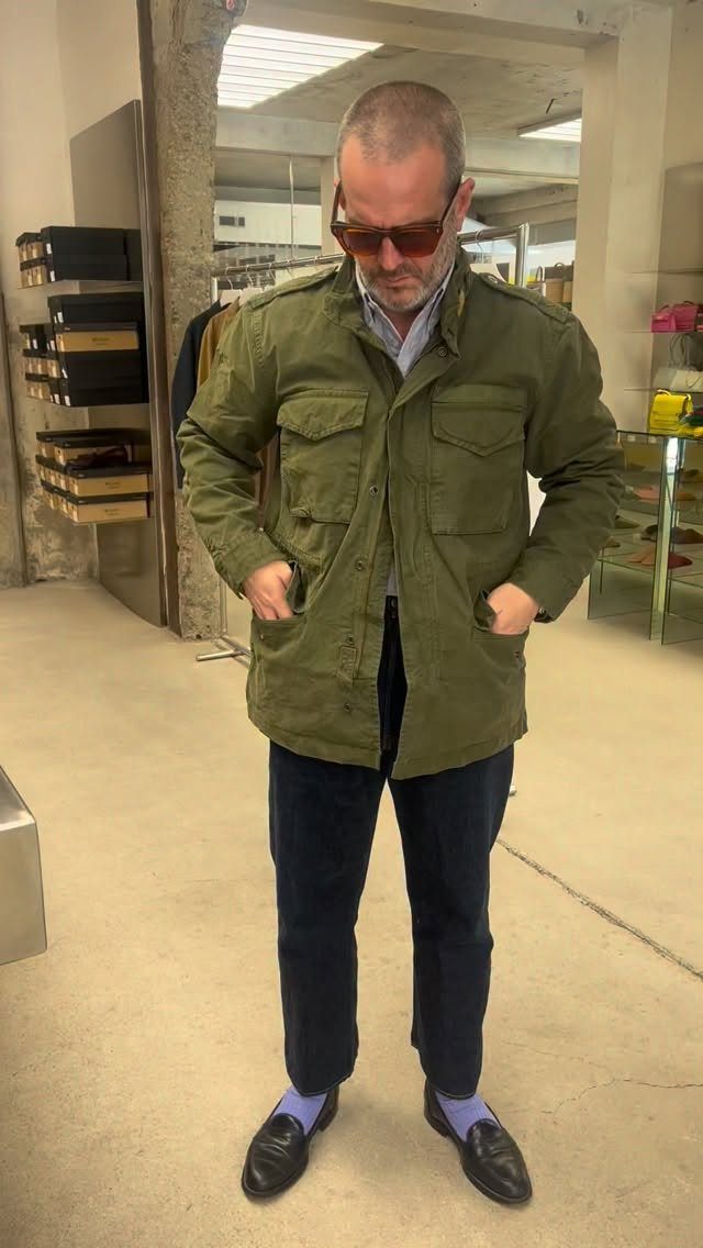 A man wearing an olive green jacket, navy trousers, and loafers with blue socks.