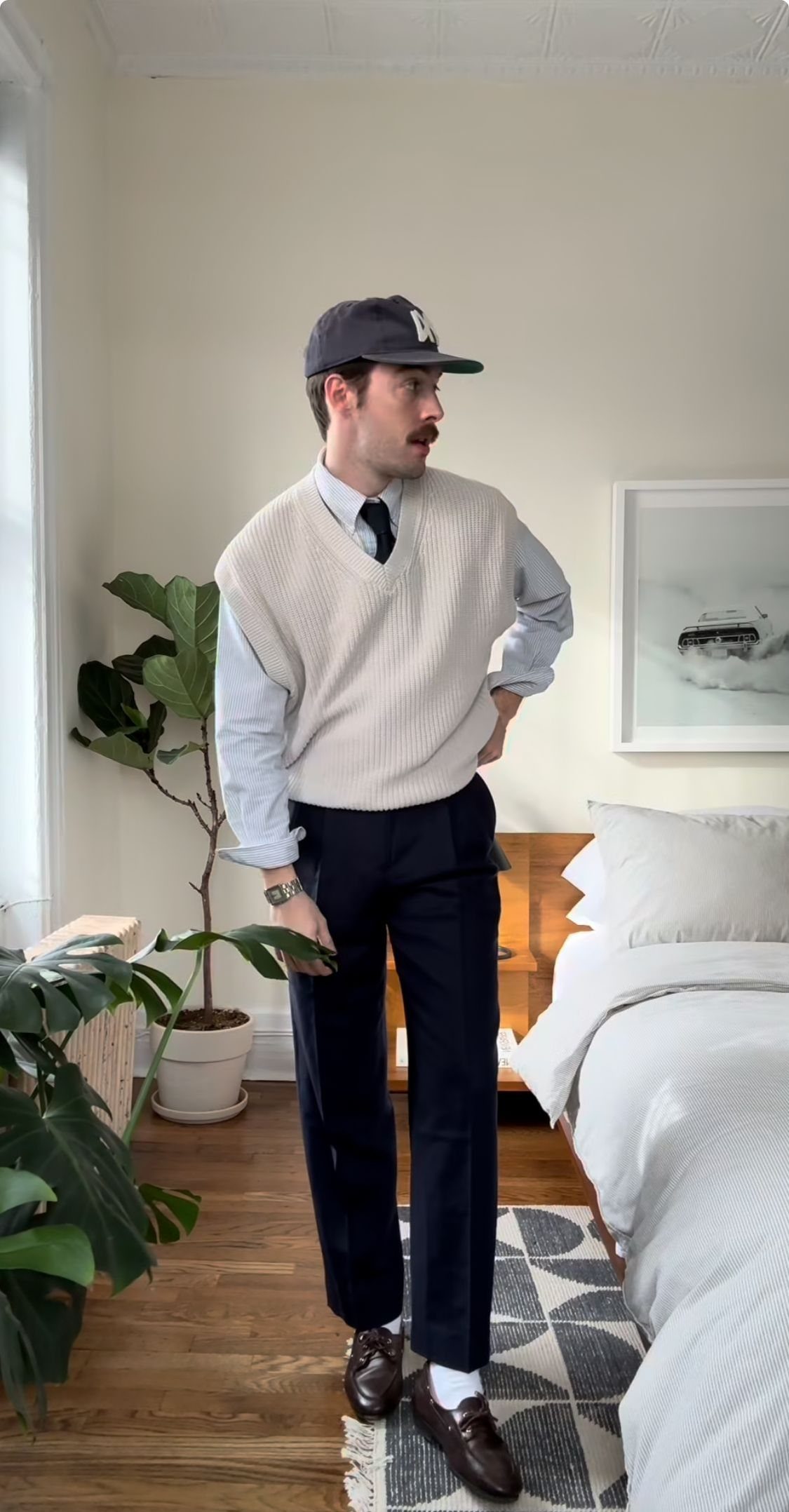 Man posing in a cream sweater vest, light blue shirt, tie, and navy trousers.