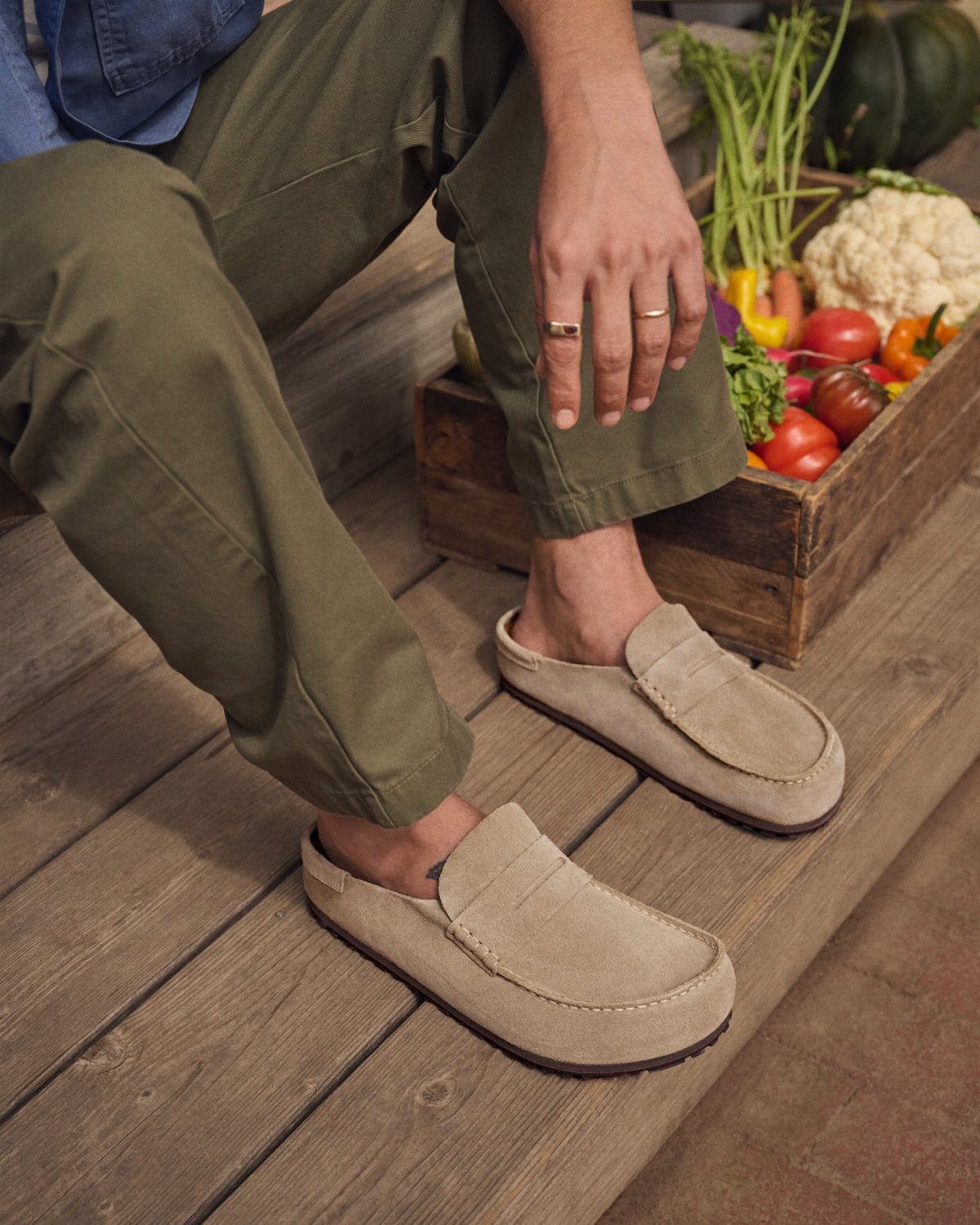 Beige suede loafers worn by a person sitting on wooden steps next to vegetables.