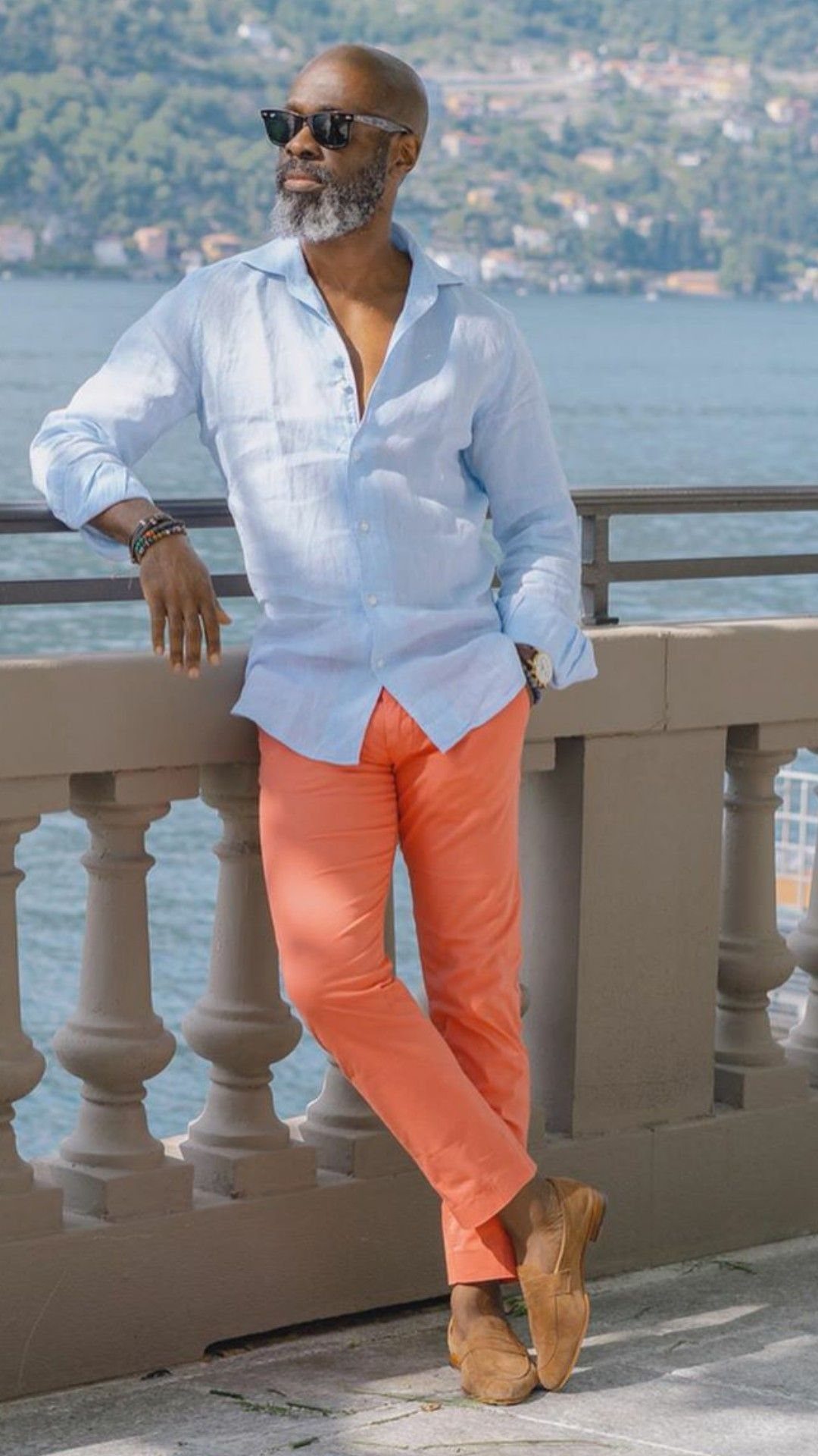 Man in a light blue linen shirt and bright coral trousers standing by a lake.