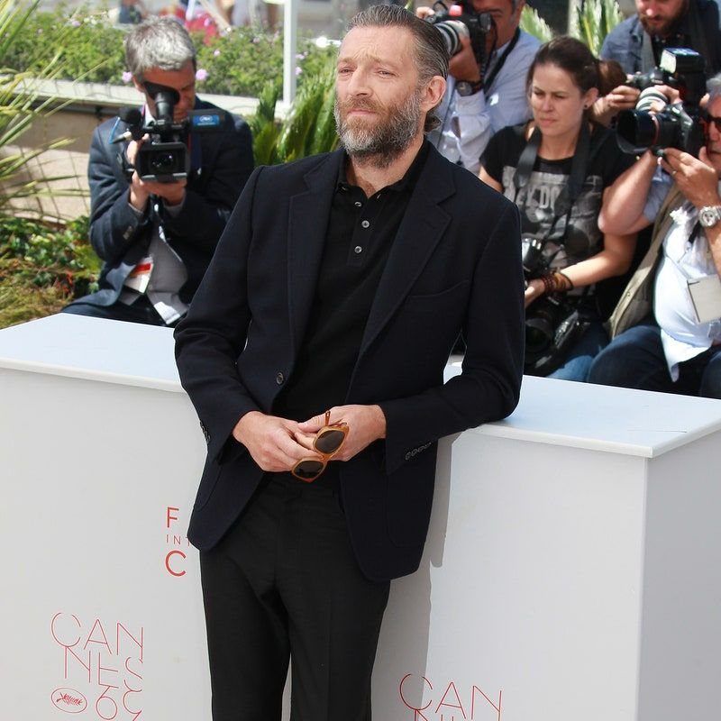 A bearded man wearing a black suit and a black polo shirt leaning against a white wall.