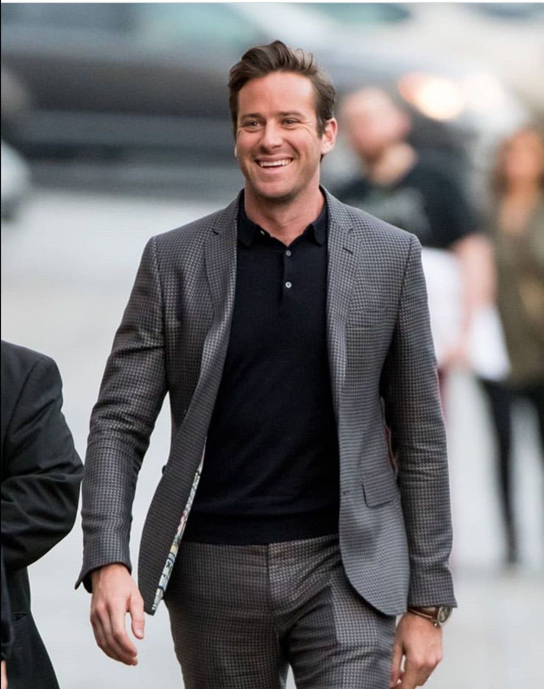 A smiling man walking outdoors wearing a grey patterned suit and a dark navy polo shirt.