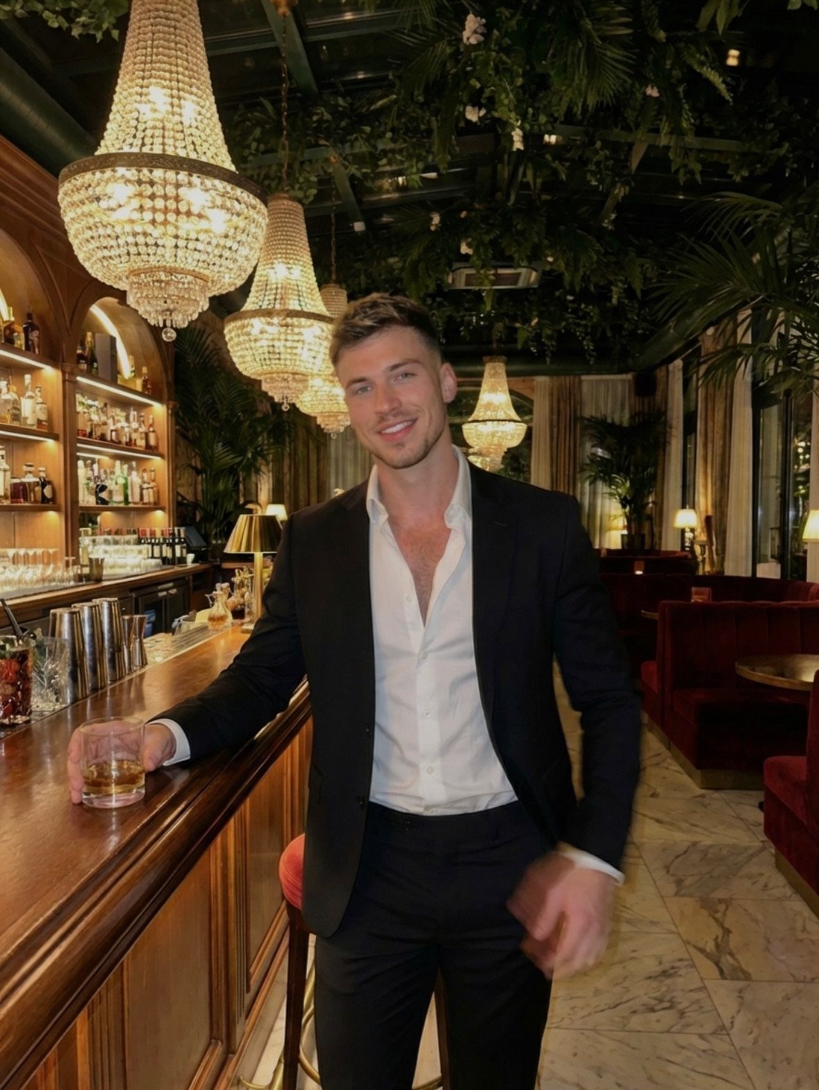 A smiling man wearing a black suit and open white shirt standing at a bar.