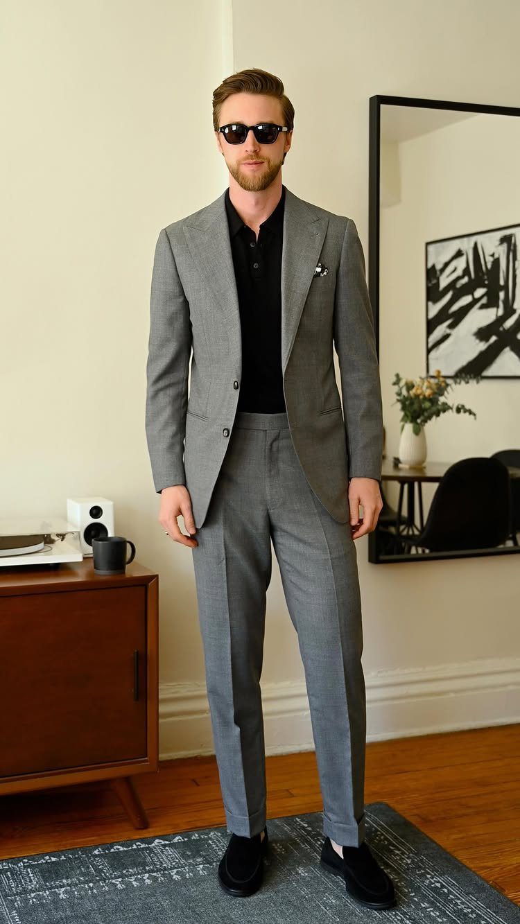 A man wearing a grey suit and black polo shirt with sunglasses standing indoors.