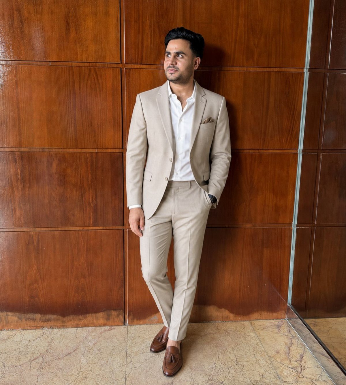 A man in a tan suit and white dress shirt posing against a wooden wall.