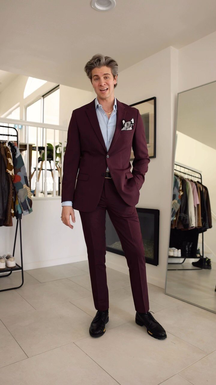 A man posing in a maroon suit and light blue open-collar shirt indoors.
