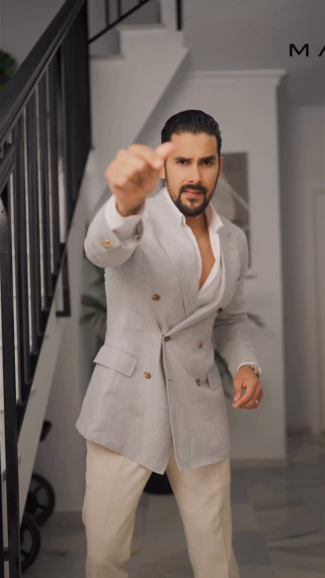 Man in a light grey linen suit with an unbuttoned white shirt pointing at camera.