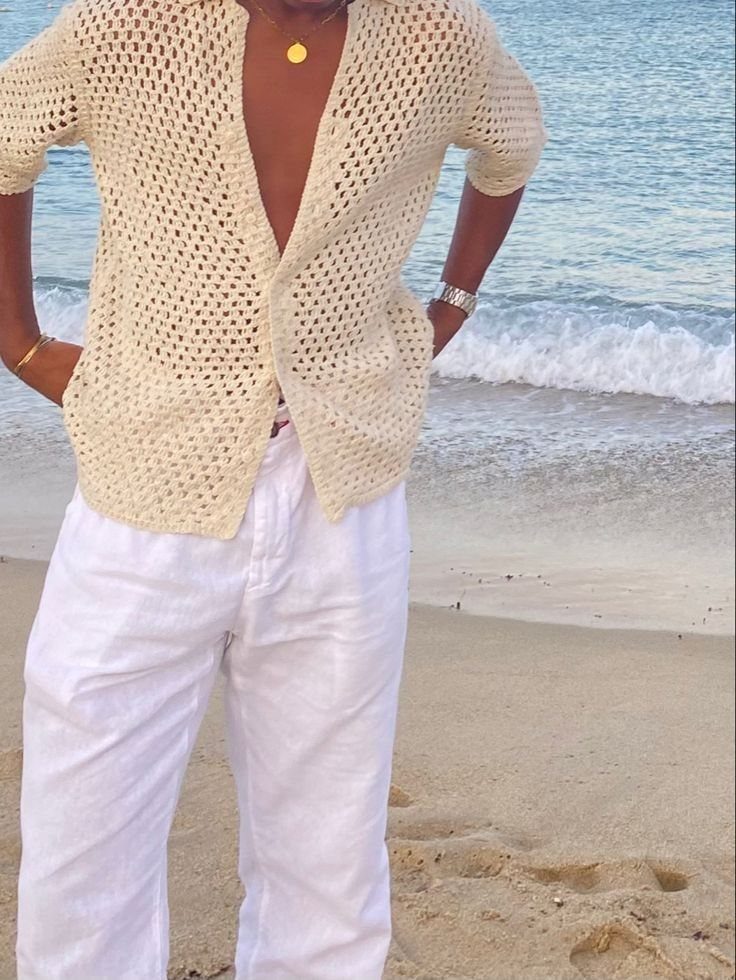 Person wearing a cream crochet shirt and white linen pants on a sandy beach.