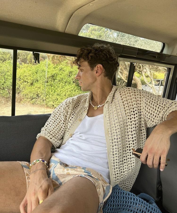 A man wearing a crochet shirt and patterned shorts sitting inside a vehicle.