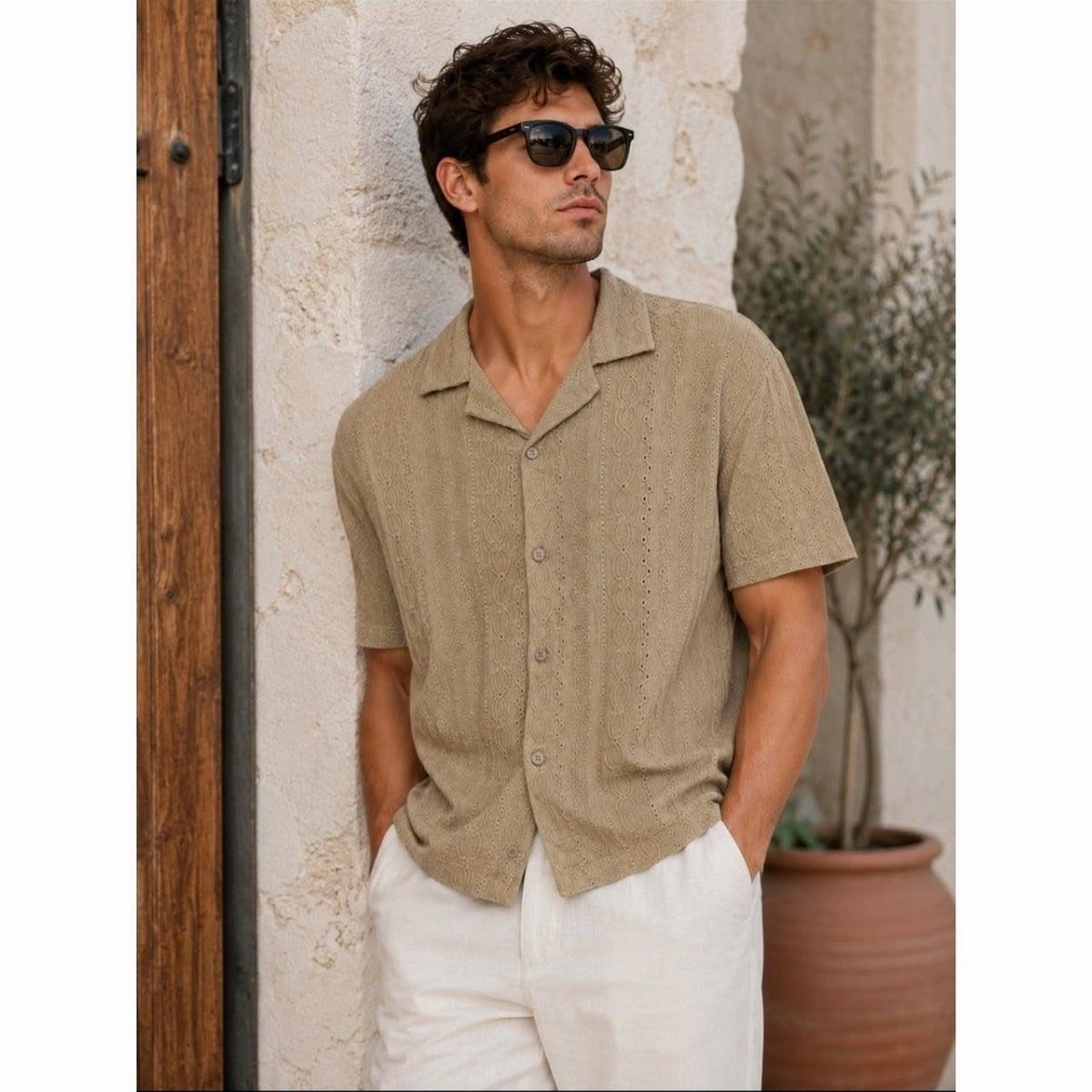 Man in tan shirt and white trousers posing against a stone wall outdoors.