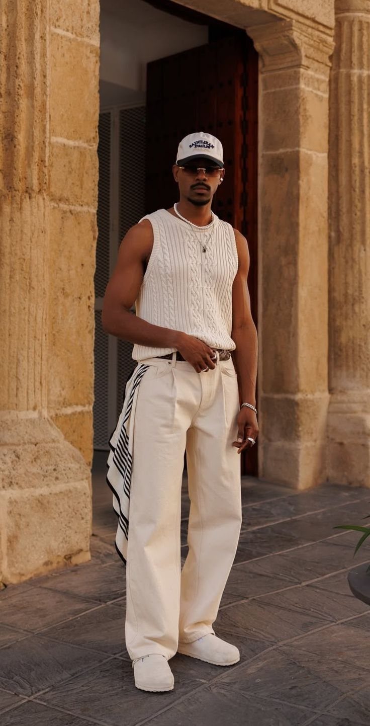 A man posing in a white knit tank top and cream trousers outdoors.
