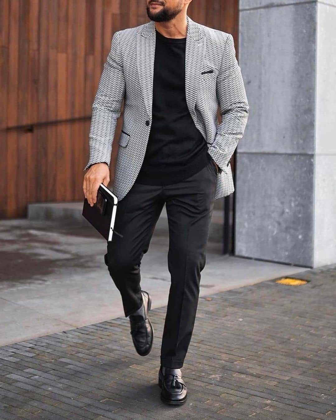 A man walking in a grey patterned blazer, black shirt, black trousers, and black loafers.
