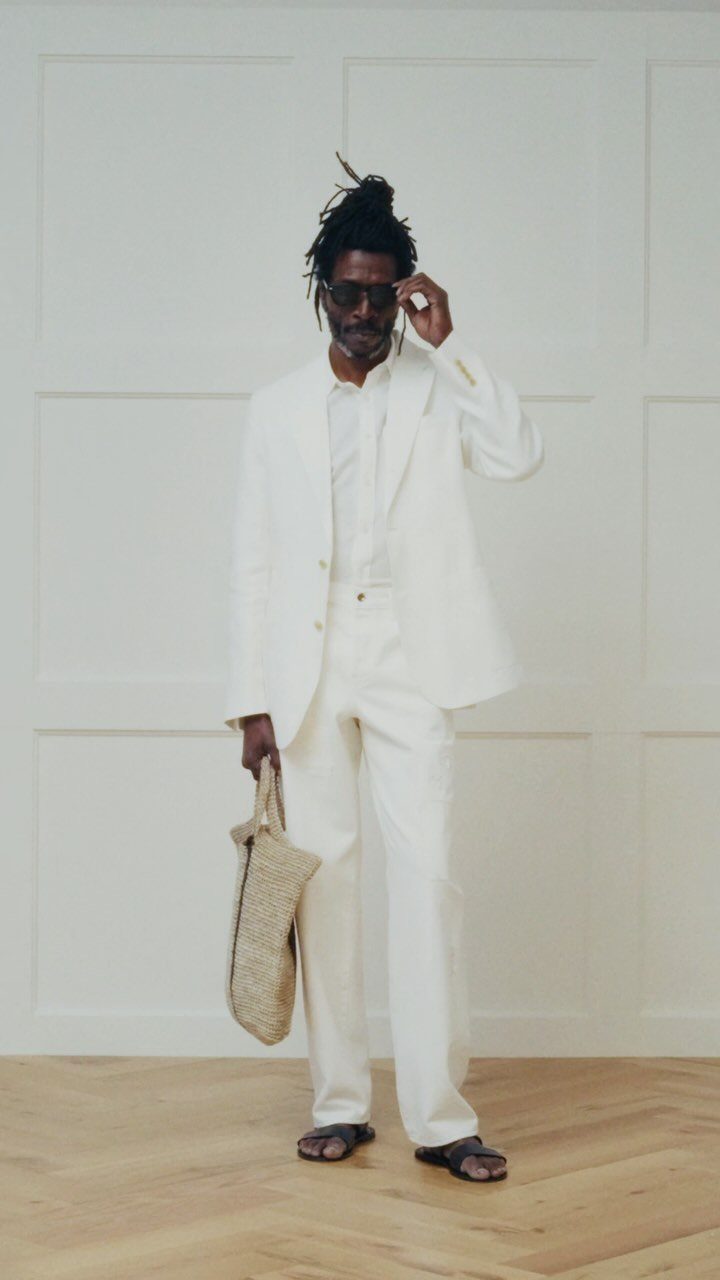 A man in a white linen suit and sandals holding a straw bag.