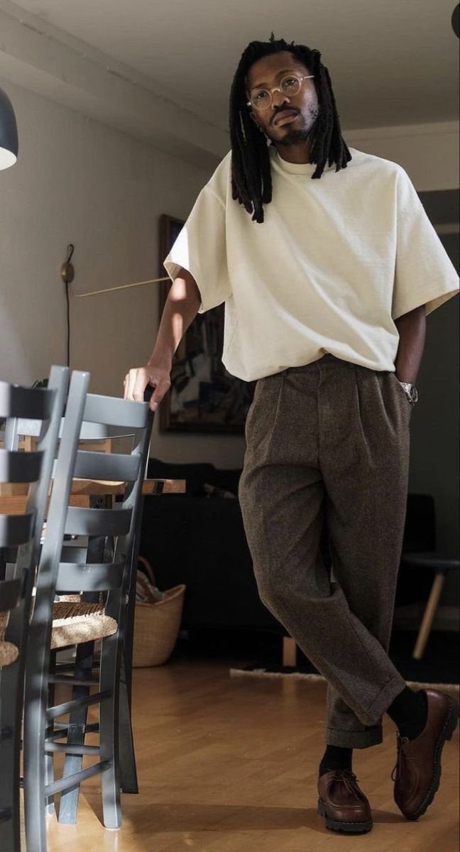 A man wearing a cream t-shirt and brown pleated trousers standing in a room.
