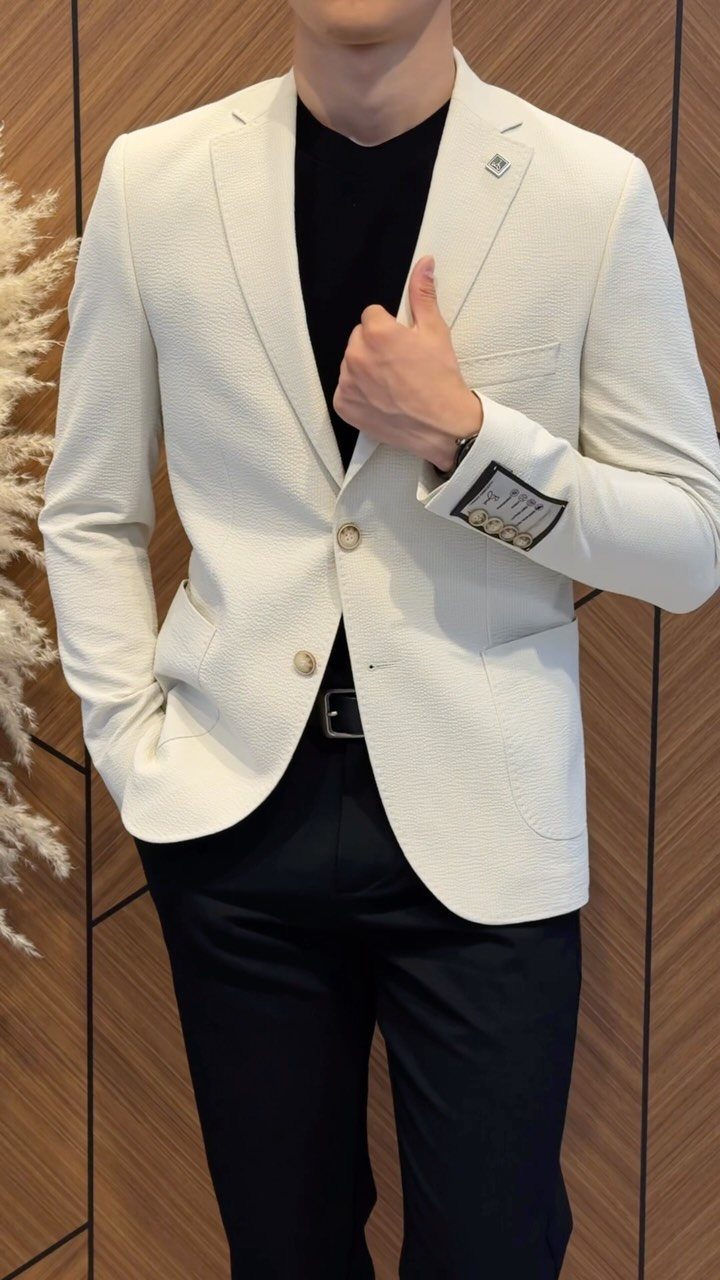 A man wearing a white textured blazer, black t-shirt, and black trousers posing against a wooden wall.