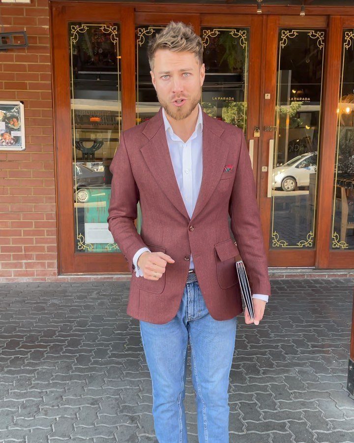 A tall man wearing a reddish-brown blazer and blue jeans standing in front of wooden doors.