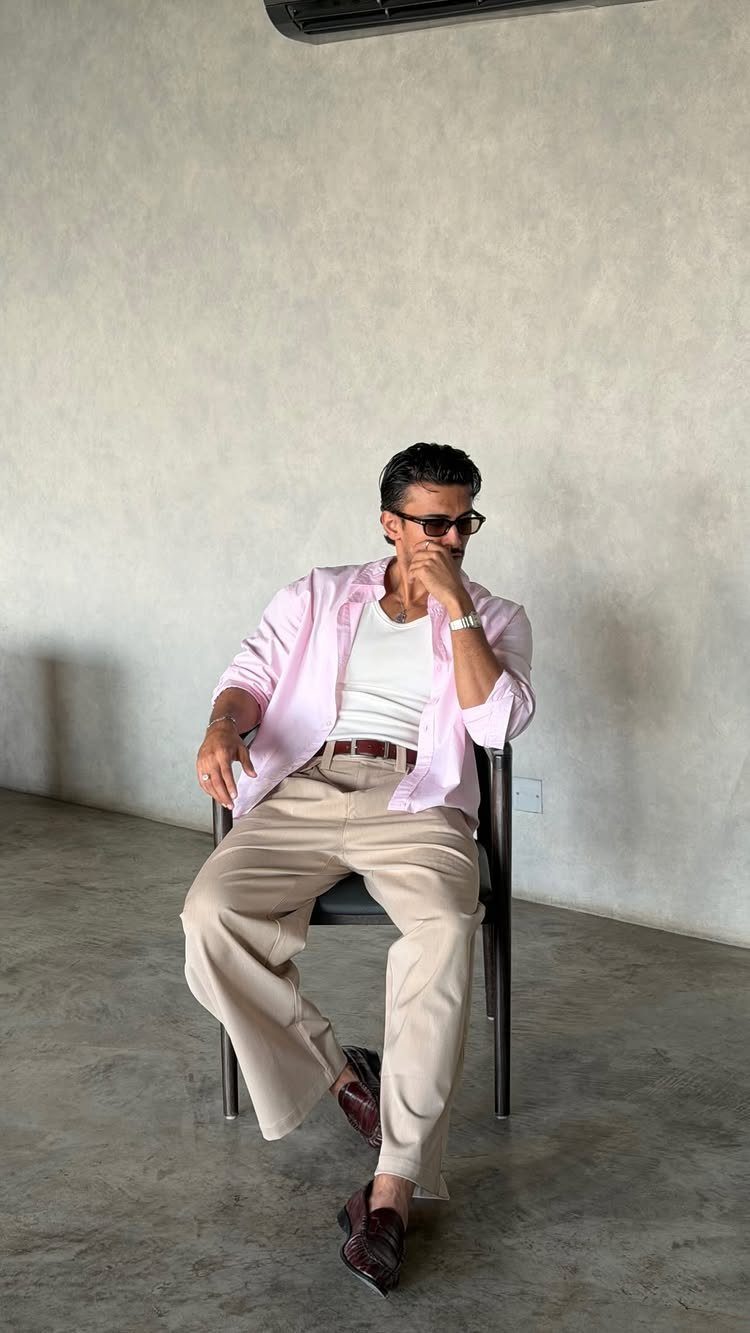 A man sitting in a chair wearing a pink shirt, white tank top, and beige trousers.