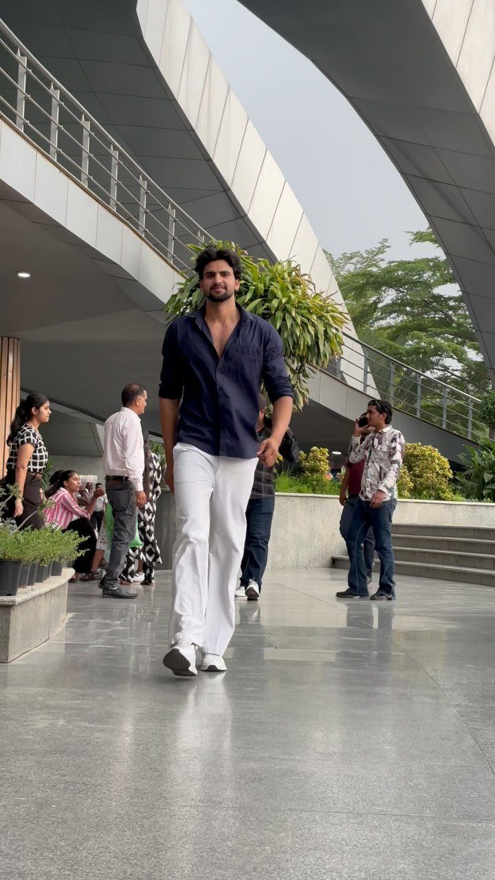 A tall man walking forward wearing a navy shirt and white trousers in a modern outdoor space.
