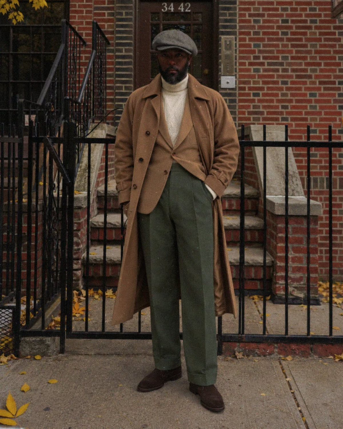 Tall man wearing a long tan overcoat, cream turtleneck, and olive trousers standing by a fence.