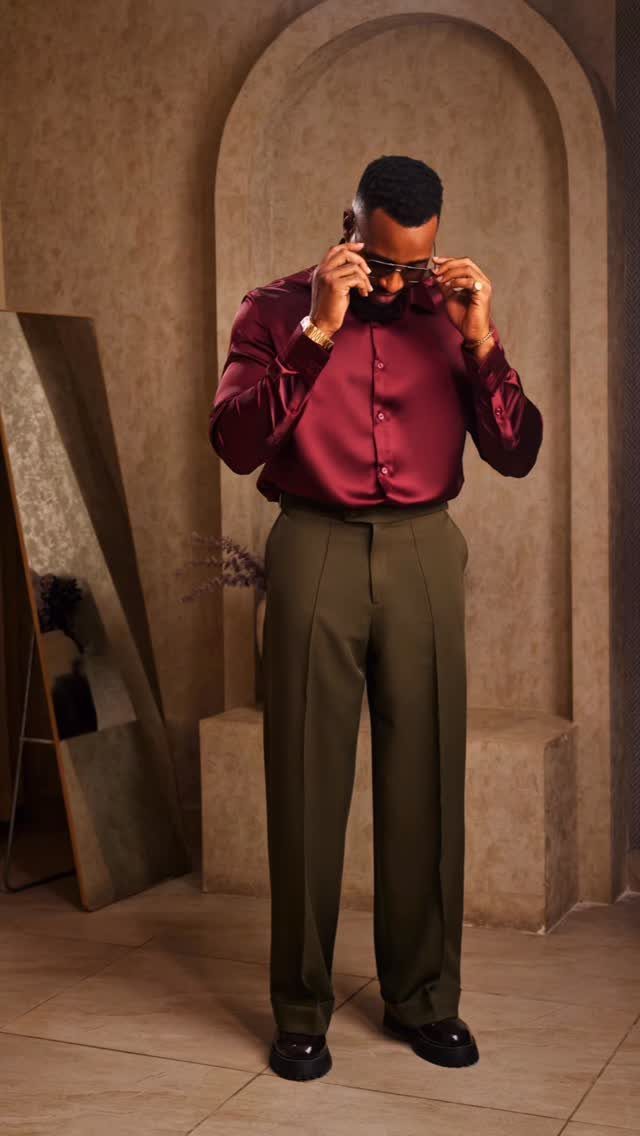 Man in maroon satin shirt and olive green pleated trousers adjusting his sunglasses.