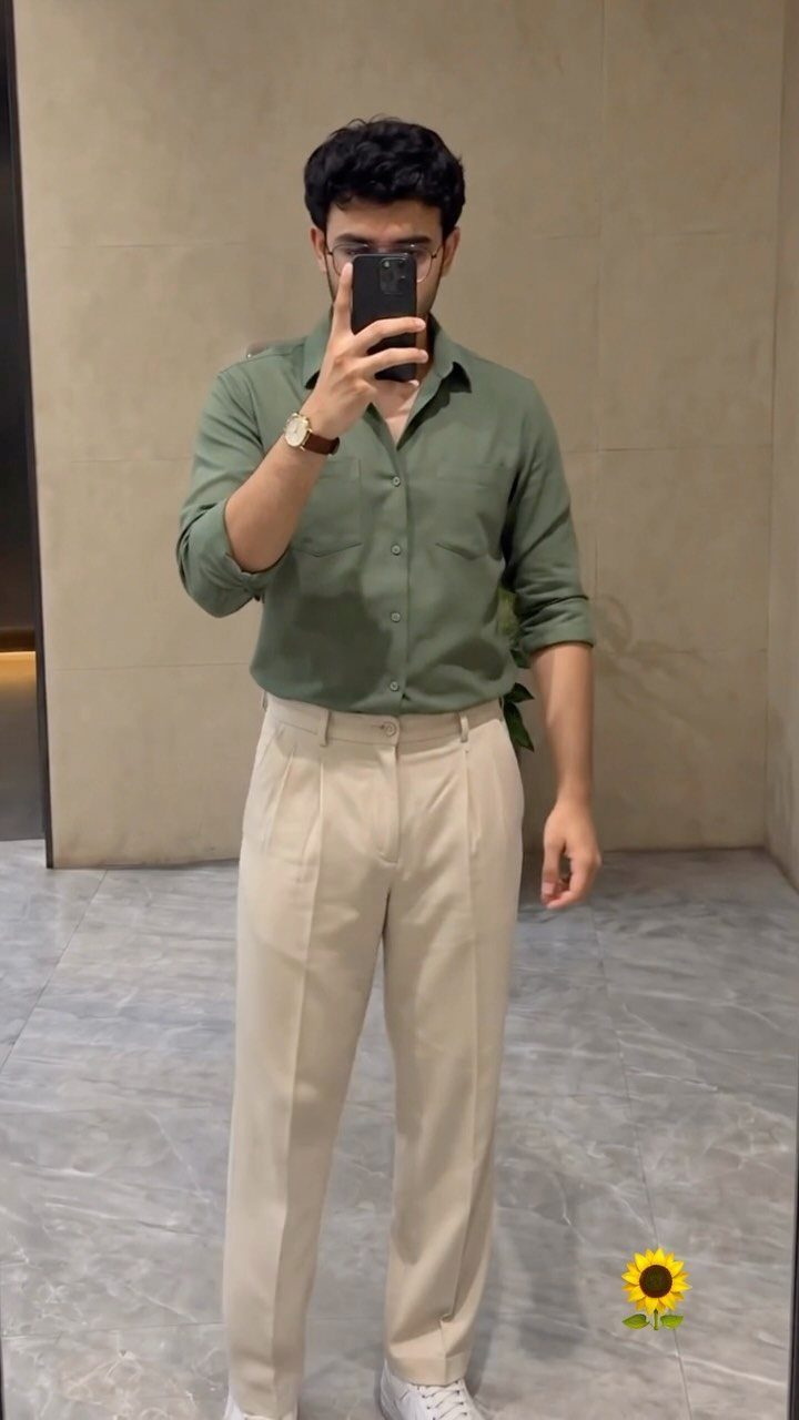 Man in a green button-down shirt and cream pleated trousers taking a mirror selfie.