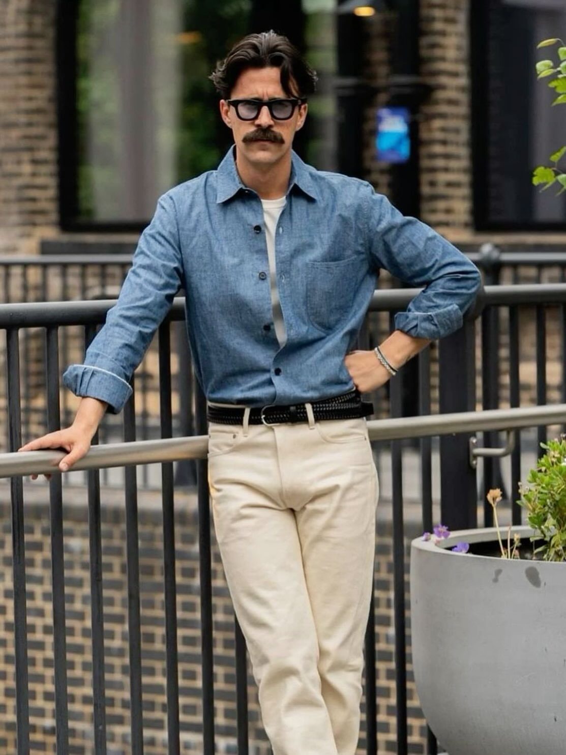 A man wearing a blue chambray shirt and cream trousers leaning against a metal railing.