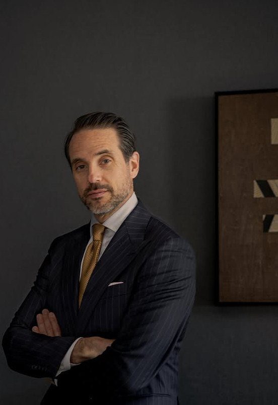 A man in a navy pinstripe suit and gold tie posing against a dark wall.