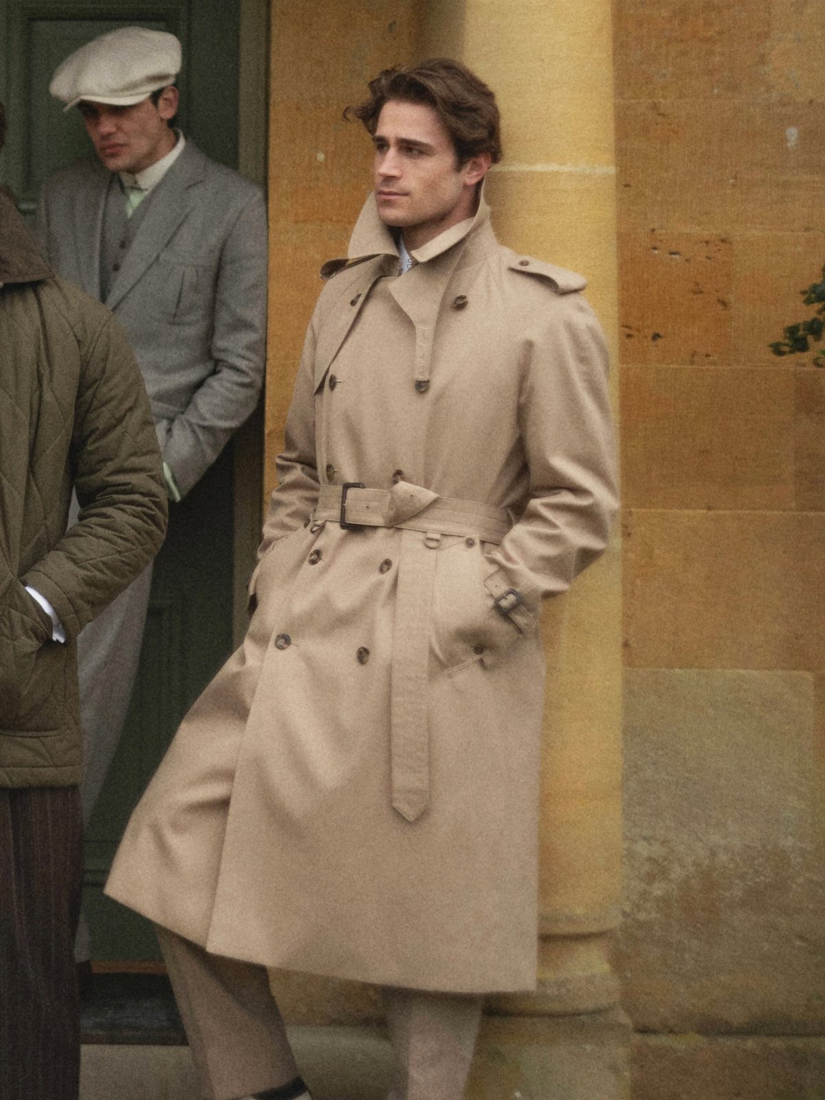 A man wearing a classic tan double-breasted trench coat standing against a stone wall.