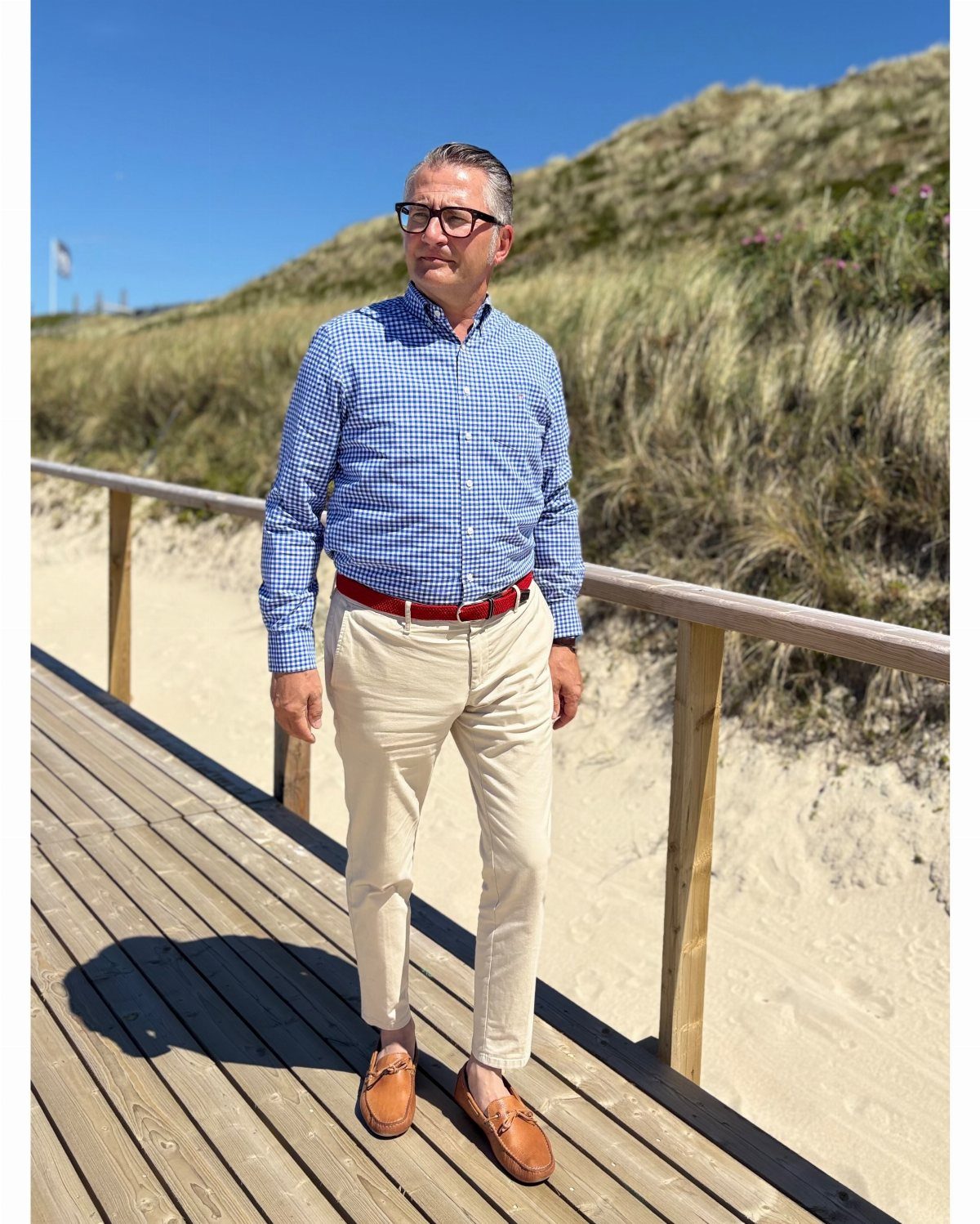 A man wearing a blue checkered shirt and beige chinos standing on a wooden boardwalk.