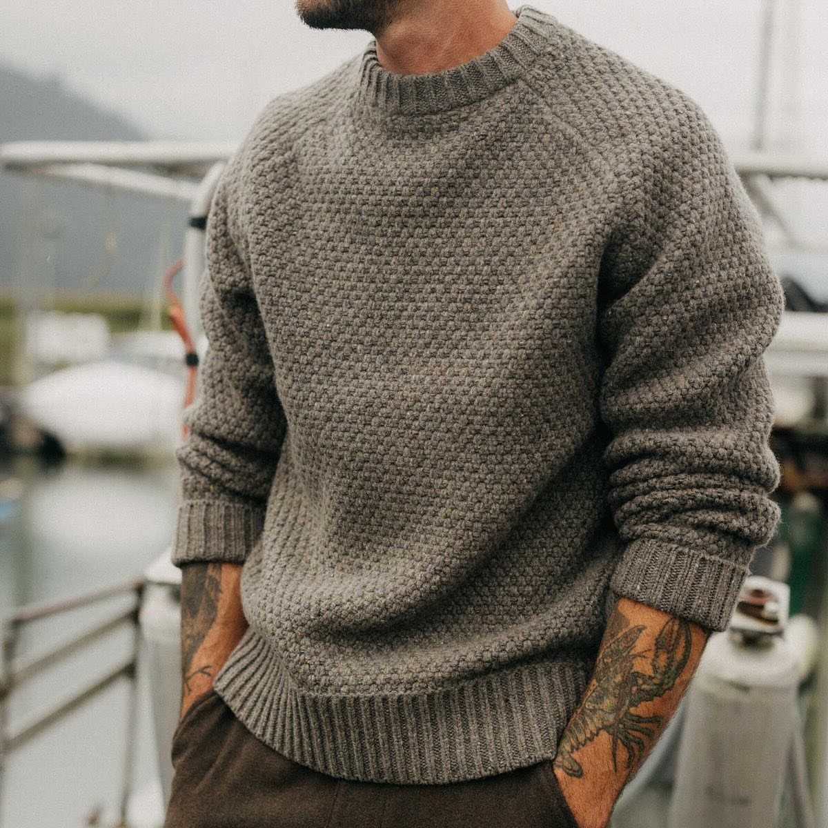 A man wearing a textured grey fisherman's sweater standing near a boat in a coastal setting.