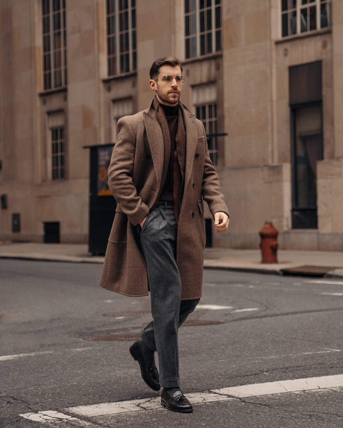 Man walking in a city wearing a long brown wool overcoat and dark trousers.