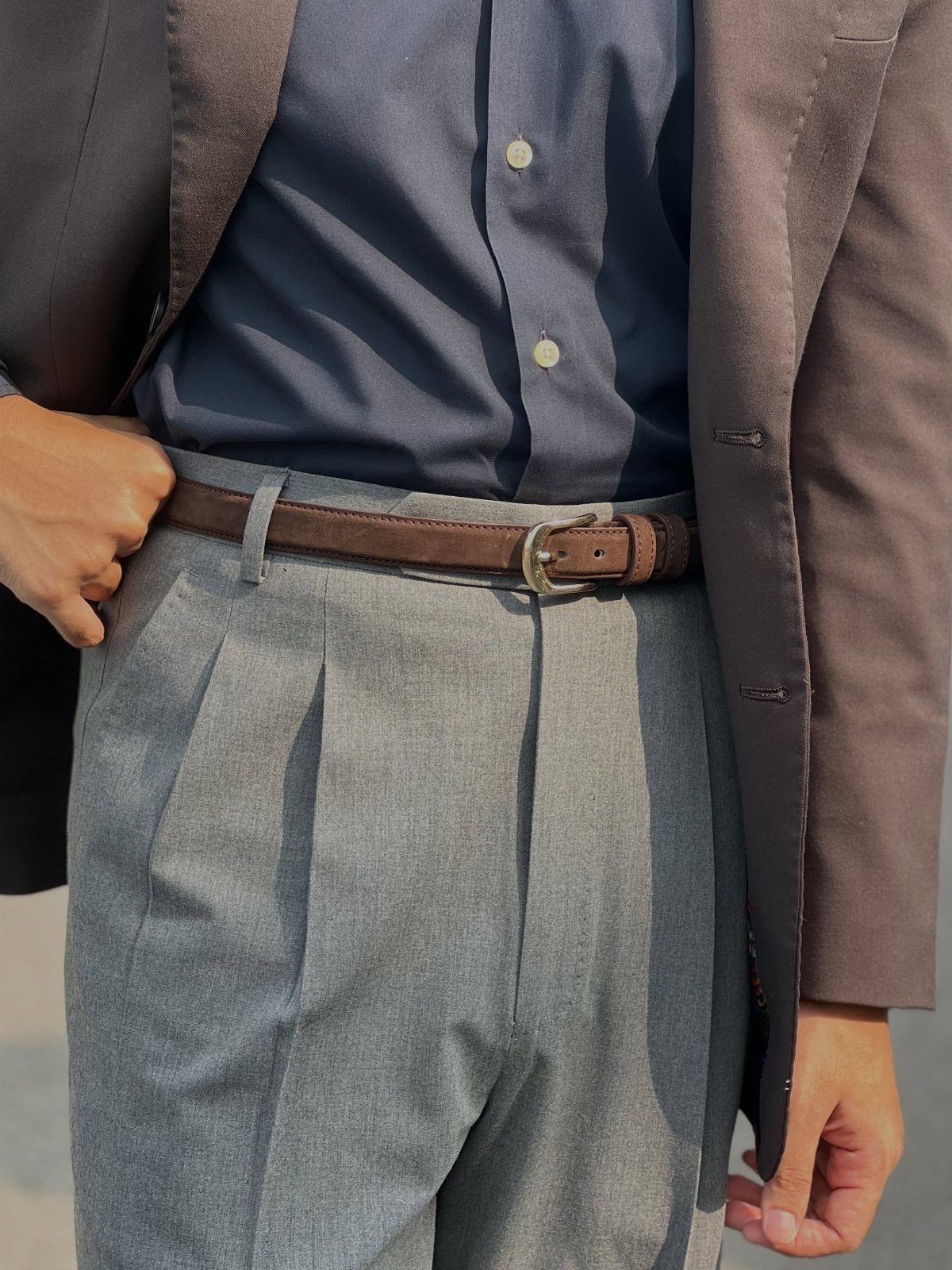 A man wearing a brown leather belt with a silver buckle and grey pleated trousers.