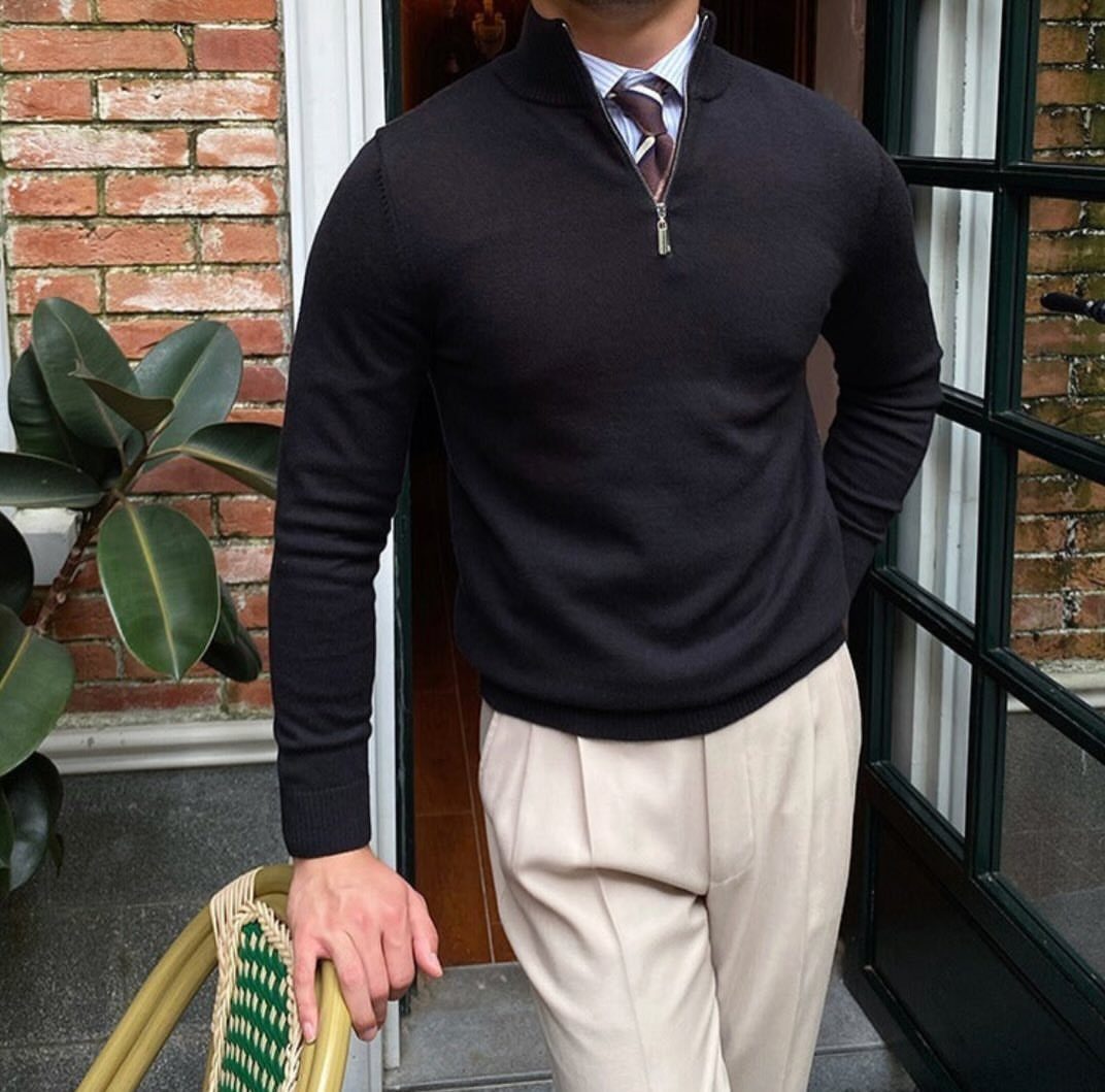 Man wearing a black quarter-zip sweater, white shirt, tie, and beige pleated trousers.