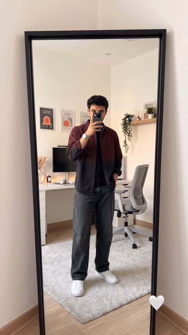A man in a dark jacket and grey jeans wearing white leather sneakers standing before a mirror.