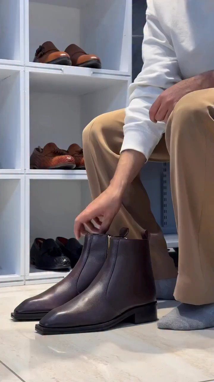 A person holding a pair of dark brown leather ankle boots near white shoe shelving.