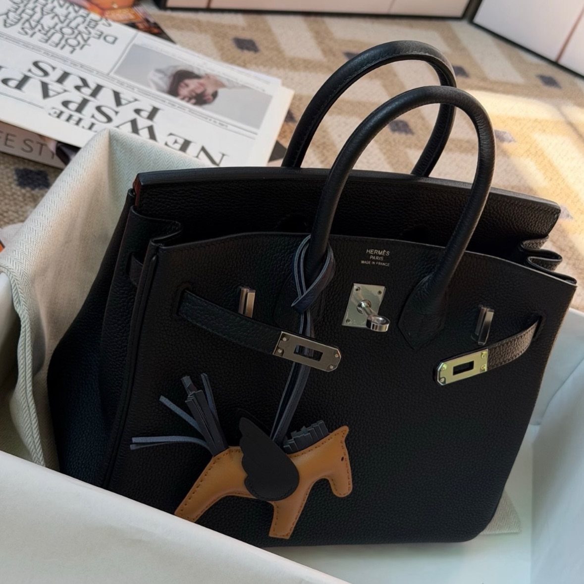 Black leather designer handbag with a horse charm resting on white fabric near a newspaper.