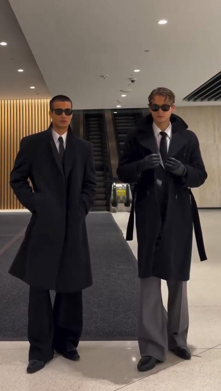 Two men wearing long black overcoats, sunglasses, and formal trousers standing in a modern building.