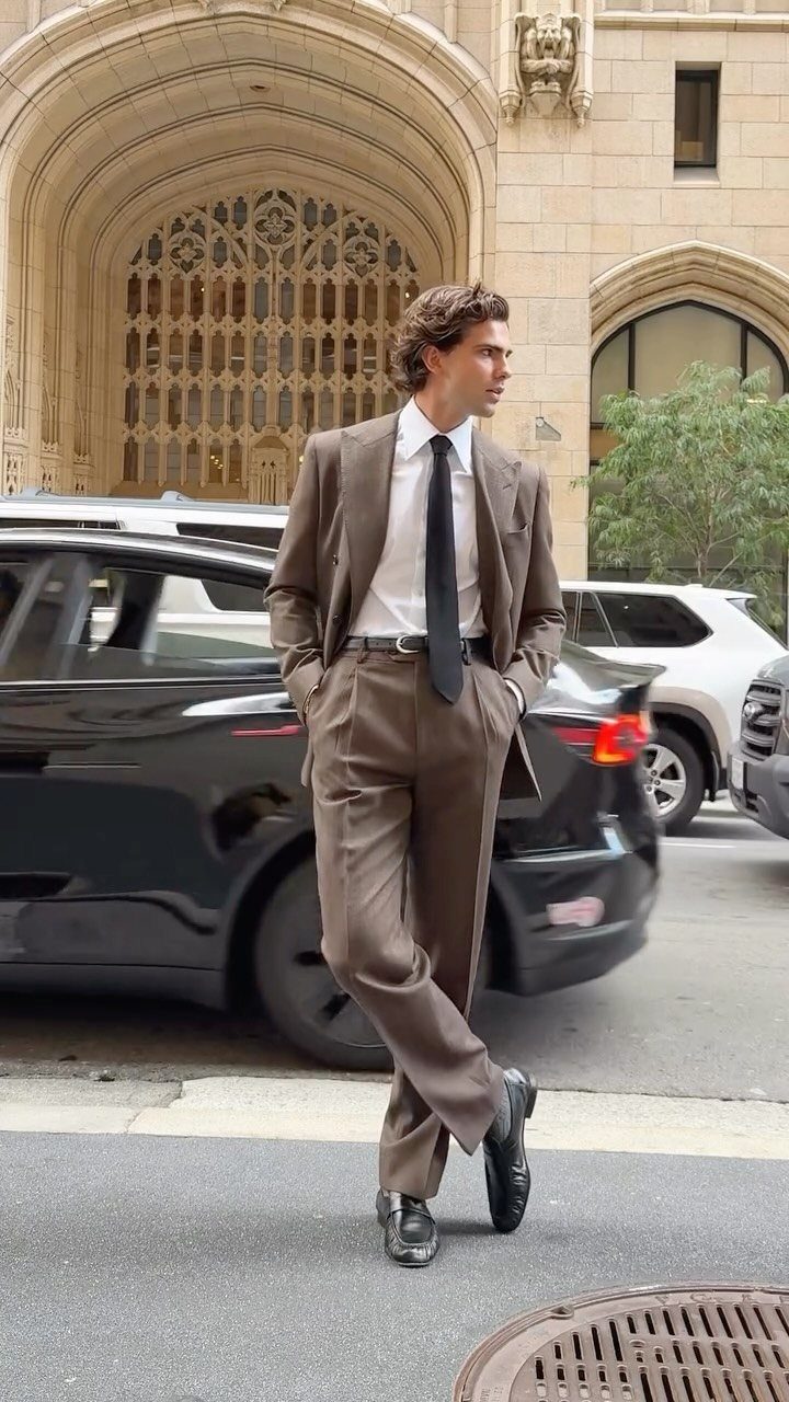 A man posing in a brown tailored suit, white shirt, and black tie on a city street.
