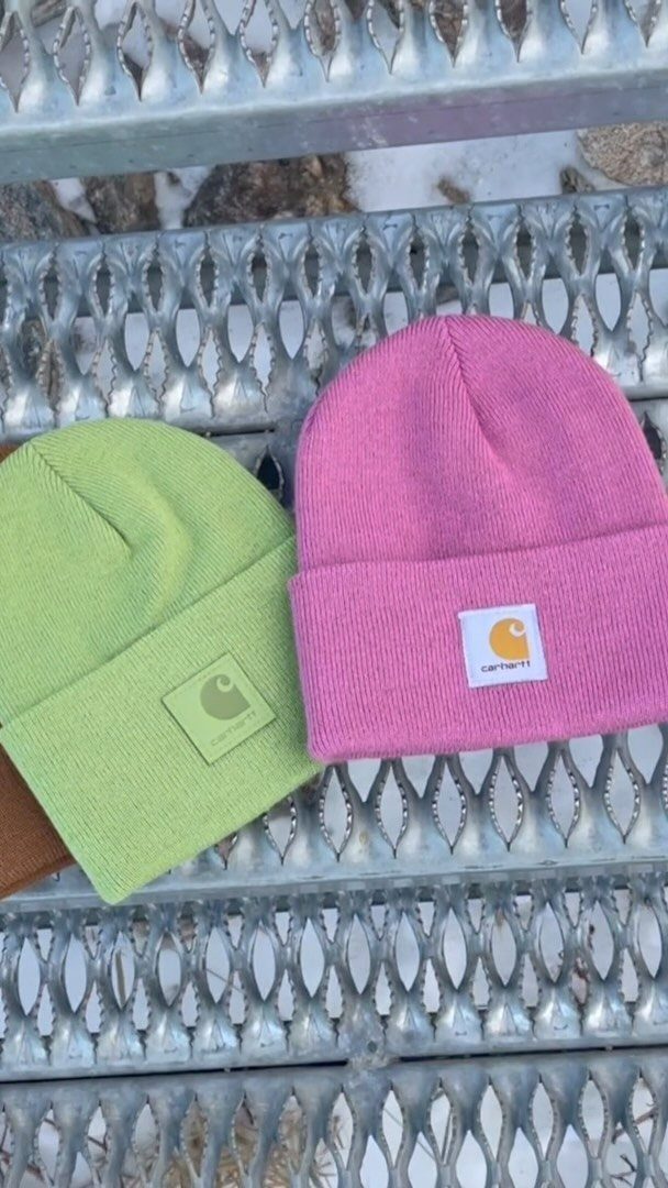 A lime green beanie and a pink beanie with Carhartt logos on a metal grate.