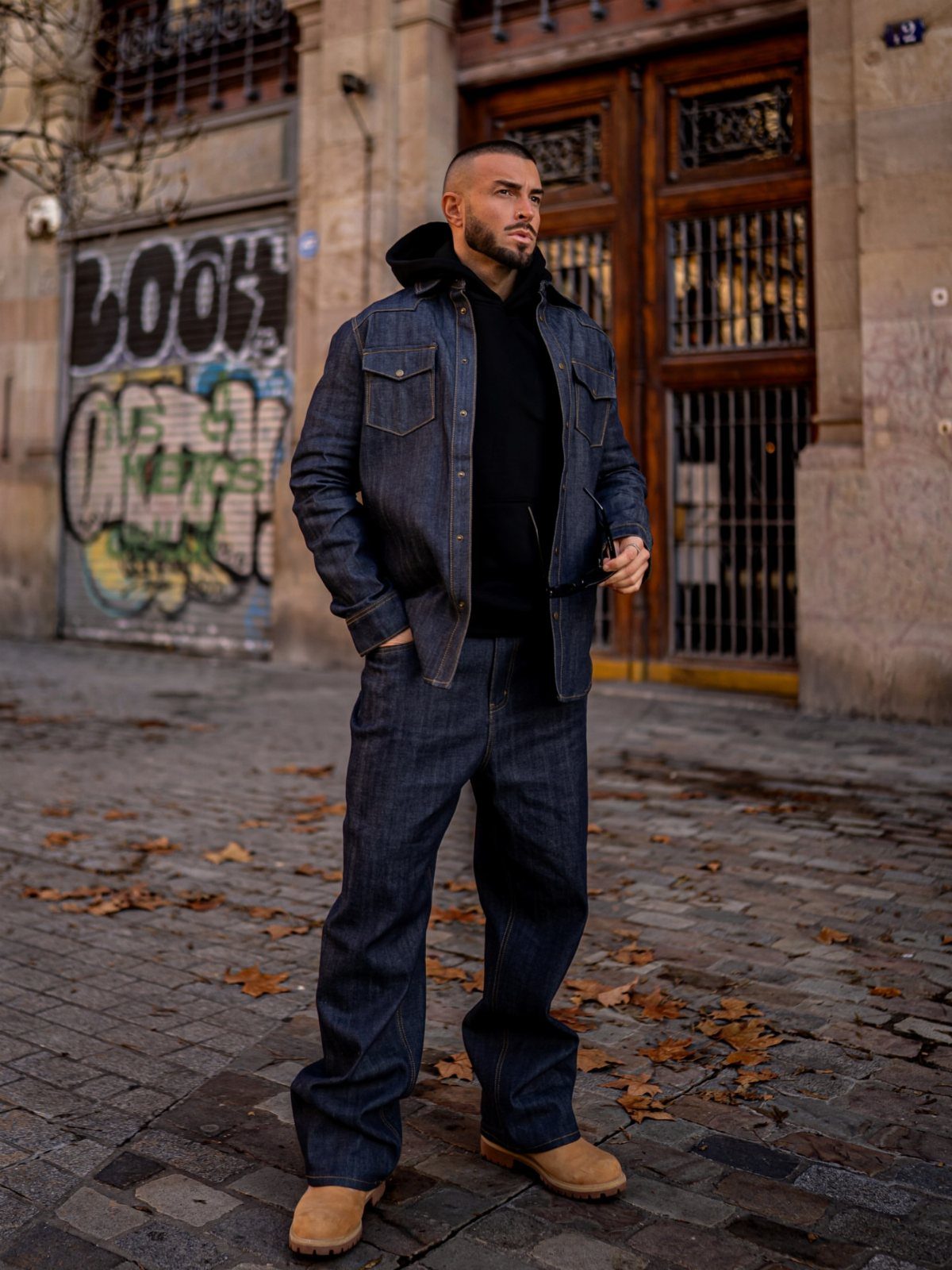 Man in dark denim jacket, black hoodie, wide denim jeans, and tan boots standing outdoors.