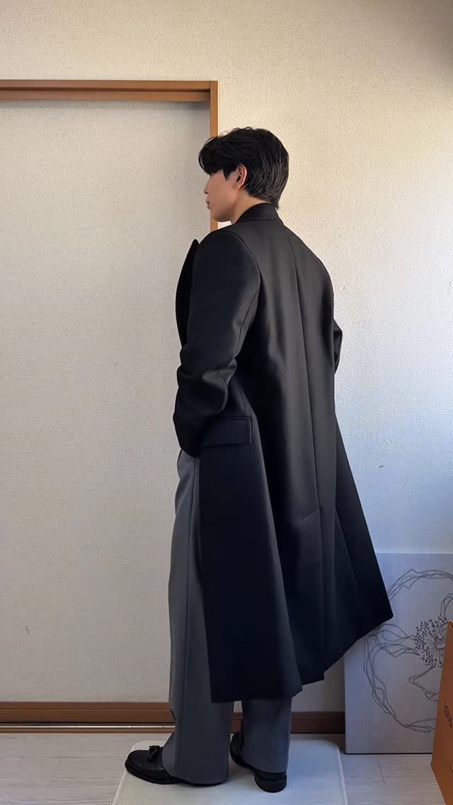 Man in a long black coat and grey trousers standing against a neutral wall.