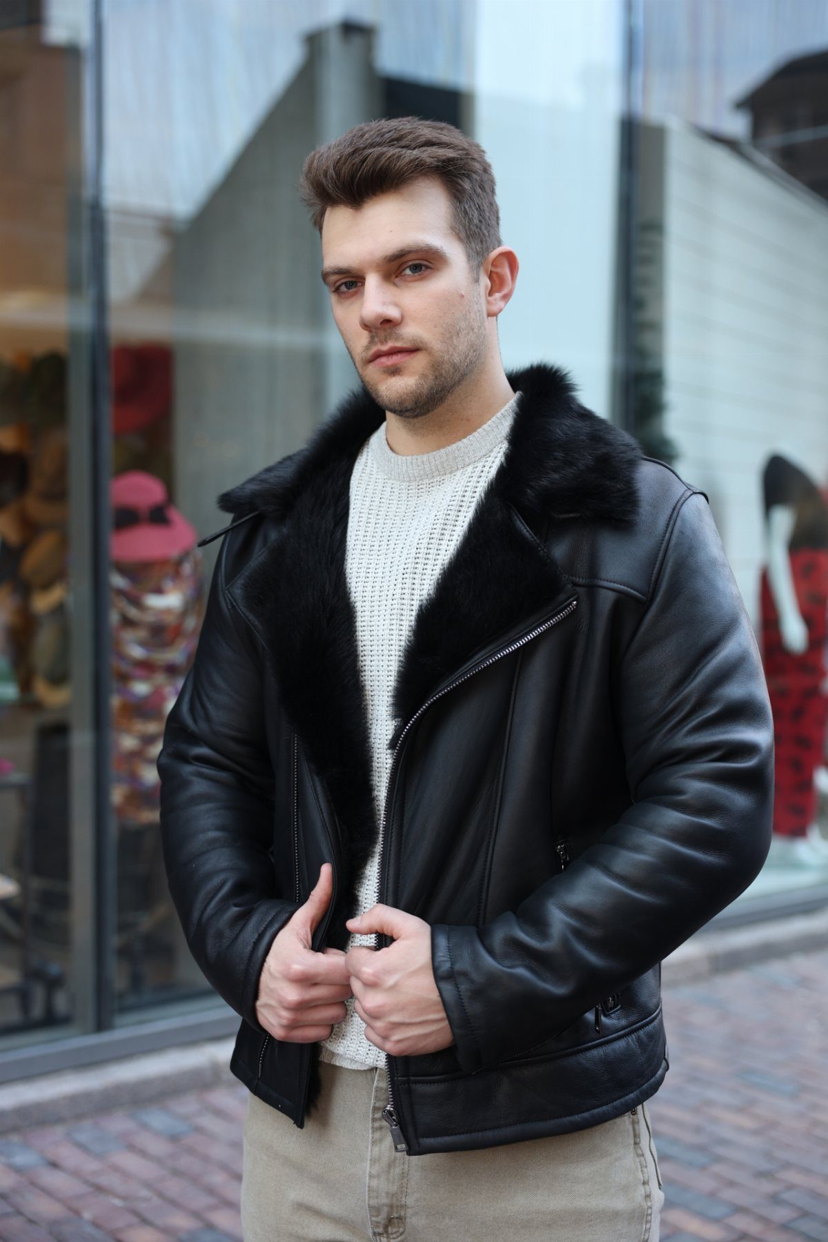 A man wearing a black leather fur-collared jacket and a white knit sweater outdoors.