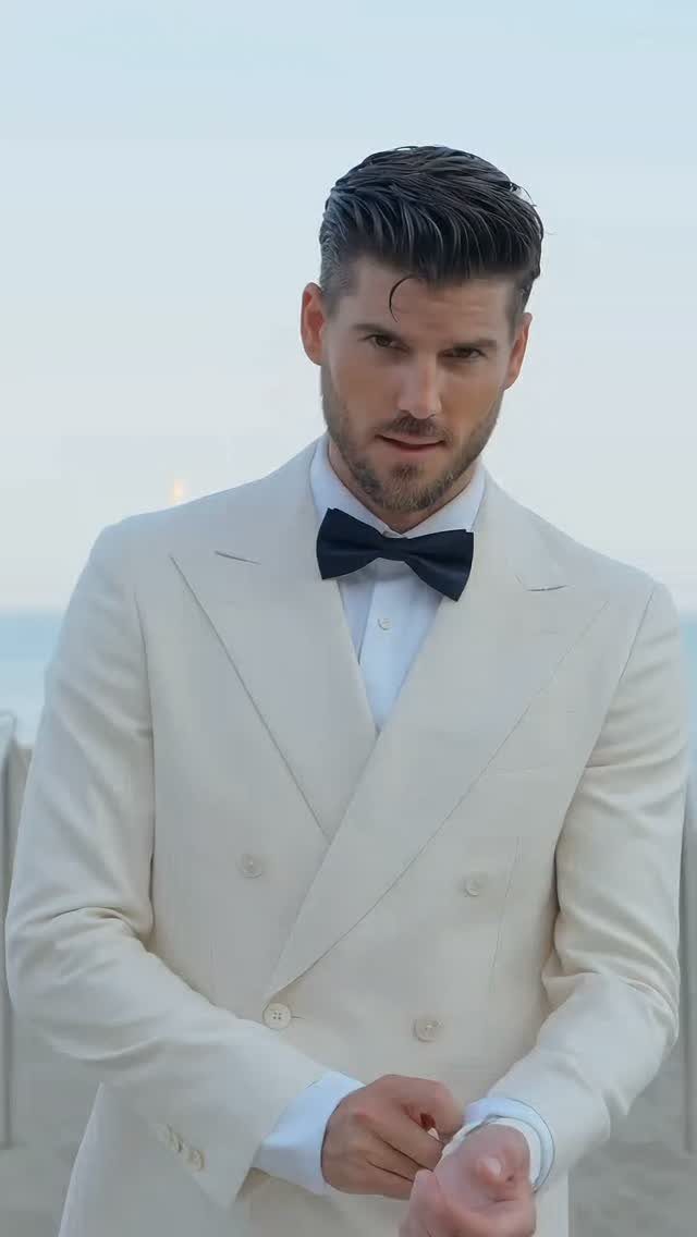 A man in a white double-breasted tuxedo jacket and black bow tie posing outdoors.